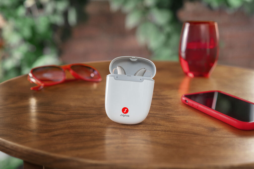 Signia MaX hearing aids in charger sitting on wooden table surrounded by red glasses, smartphone with red case
